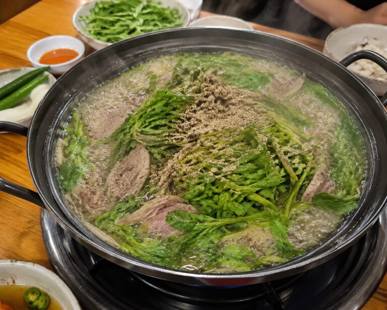 담양 오리 맛집 유진정의 청둥오리전골이 끓는 모습으로 솥 안에 오리고기와 미나리, 들깨가루가 보이며 주변에 밑반찬과 소스가 세팅되어 있다.