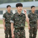 페이커 이상혁이 군복을 입고 기초군사훈련을 받으며 미소 짓는 모습, 배경은 육군훈련소 내 야외 훈련장
